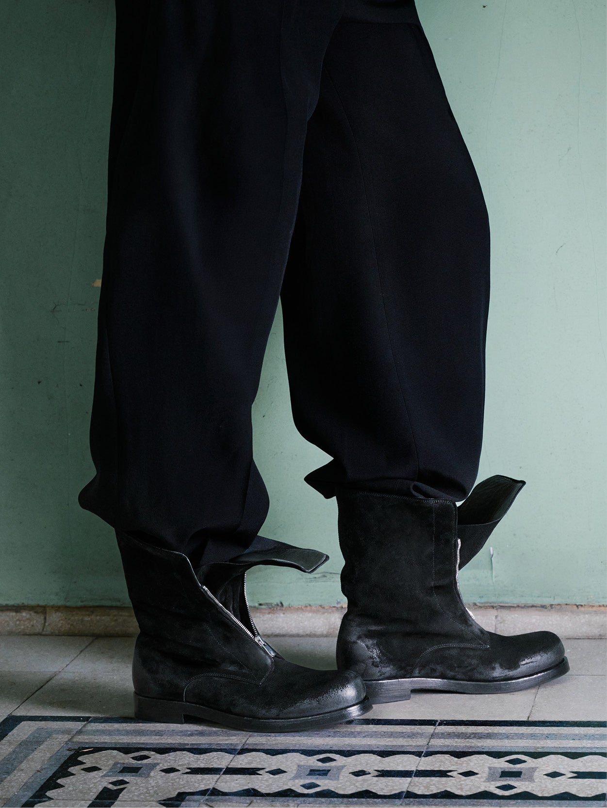 A person wearing loose black pants and black leather ankle boots with side zippers, standing indoors against a pale green wall.
